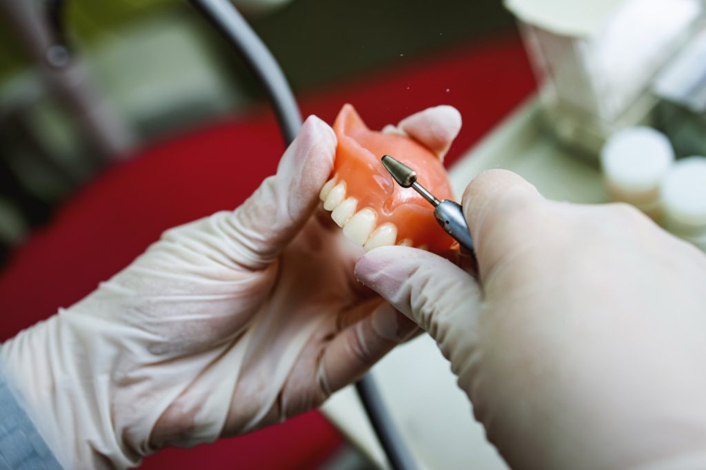Close-up of a dental technician shaping a prosthesis tooth with a tool at work desk.
